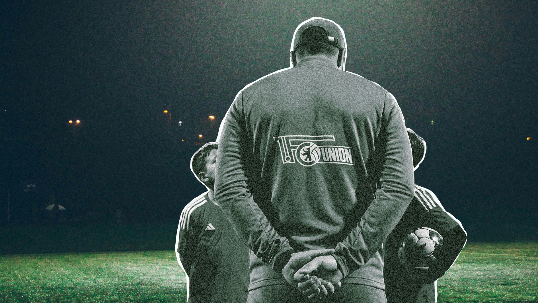 Fußballtrainer steht vor zwei Fußballspielern. Auf dem Rücken des Trainers ist ein Logo des Fußballvereins Union Berlin zu sehen.