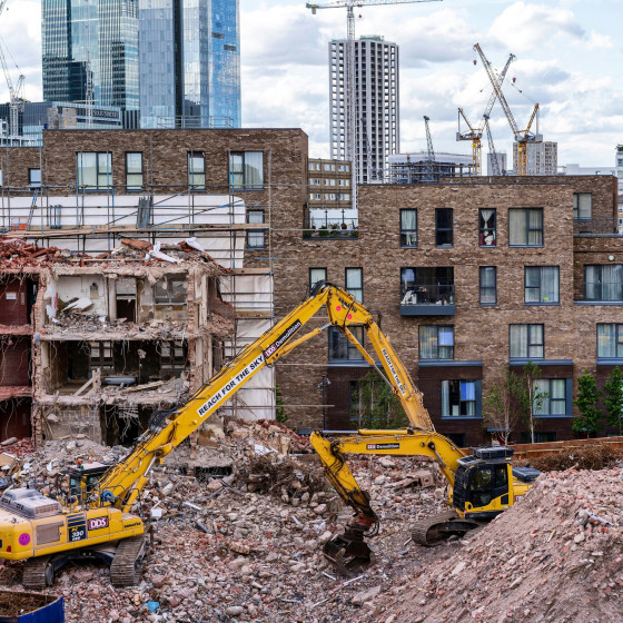Ein Bagger steht auf einem Haufen Bauschutt, dahinter ein zur Hälfte abgerissenes Haus und viele moderne Neubauten